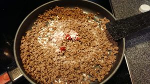 A frying pan with browned minced meat. In it is some red splotches of tomato puree and white splotches of cream.