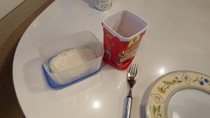 A container with shredded coconut and a box of raisins next to a fork and a plate on a white table.