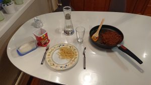 A white table with a plate with spaghetti on it, a fork to the left, a knife to the right, a glass to the upper right, a frying pan with minced meat and tomato- and cream sauce, a carafe with water, a box of raisins, and a container with shredded coconut.