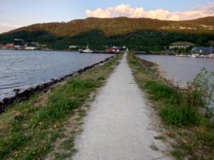 En smal sti på en molo ut mot et hvitt bygg i enden. På venstre side, fjorden. På høyre side, småbåthavn.