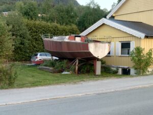En gammel, slitt båt står støttet opp ved siden av et lysegult hus.