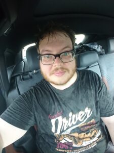 En mann med briller, frisert skjegg, og sort t-skjorte. Han er gjennomvåt og sitter i baksetet på en bil.