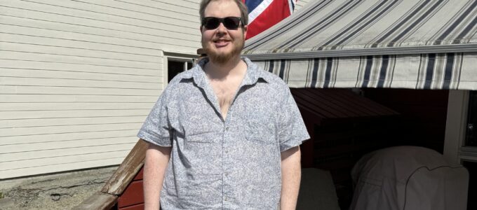En skjeggete mann med solbriller står og smiler på en terrasse. Han har på seg en beige, knelang shorts og en grå, kortermet skjorte med hvitt mønster. I bakgrunnen, en stol, en markisa, og det norske flagget.