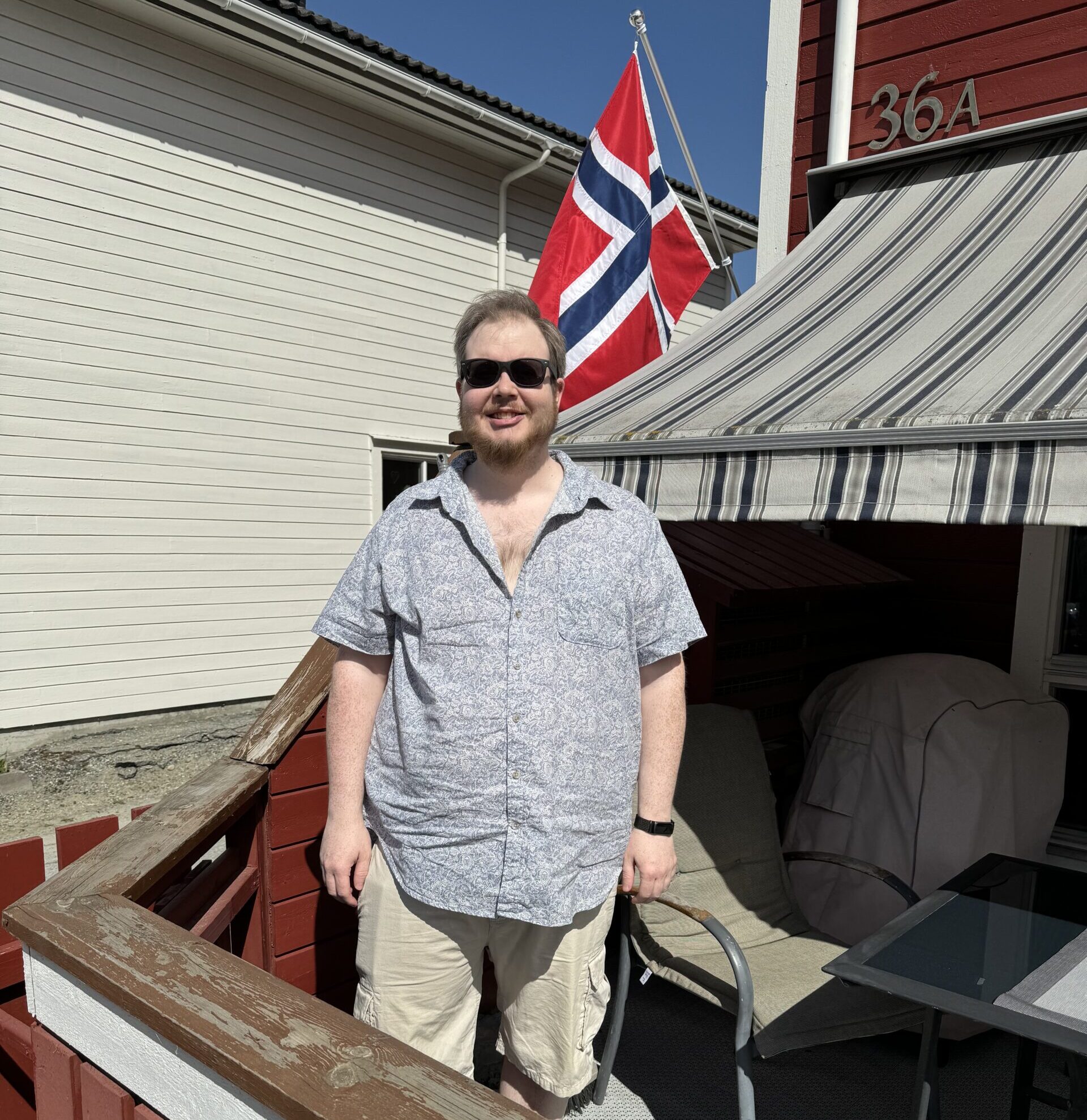 En skjeggete mann med solbriller står og smiler på en terrasse. Han har på seg en beige, knelang shorts og en grå, kortermet skjorte med hvitt mønster. I bakgrunnen, en stol, en markisa, og det norske flagget.