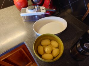 Pealed potatoes in a tupperware, a white ovenproof dish, and a white mandoline.