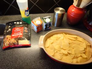 A bag of grated cheese, two boxes of cream, salt, and an ovenproof dish with sliced potatoes in it.