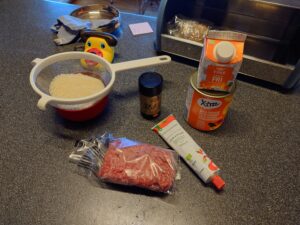 A colander with rice, a bag with minced meat, a box of spice mix (Natvigs krydderblanding), a tube of tomato puree, a can of halved peaches, a box of lactose free heavy cream.
